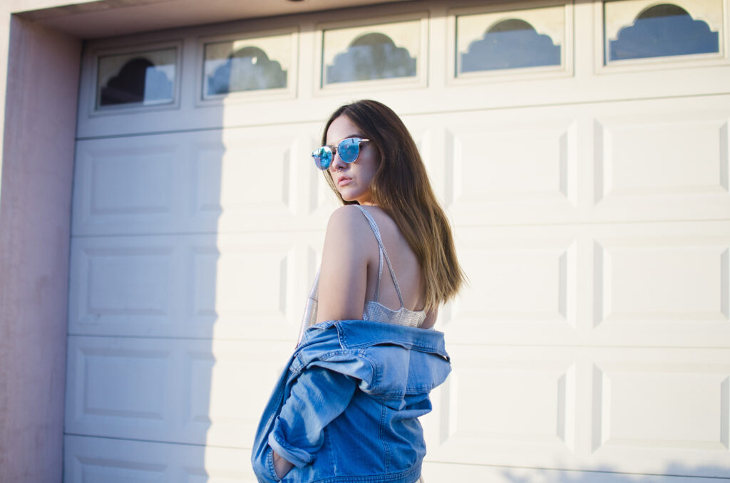 Laura Adriana photo shoot in front of a pink garage door with blue sunglasses and a denim jacket.